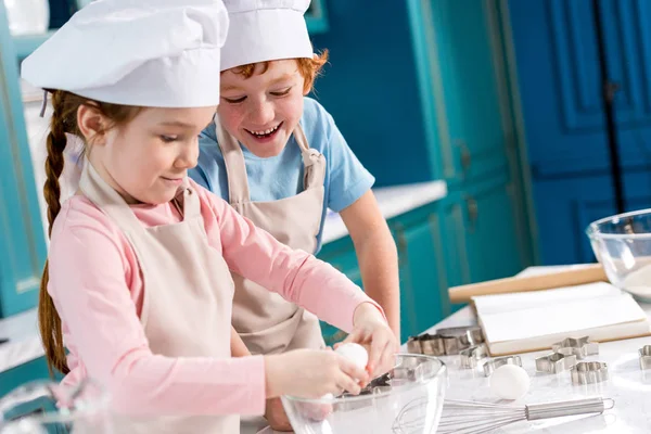 Adorables niños felices en sombreros de chef y delantales haciendo masa juntos en la cocina - foto de stock