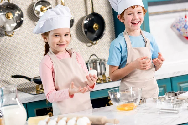 Adorables niños felices en gorros de chef y delantales sonriendo a la cámara mientras hacen masa juntos en la cocina - foto de stock