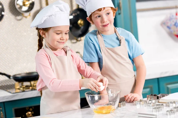 Niedliche kleine Geschwister in Kochmützen und Schürzen bereiten gemeinsam in der Küche Teig zu — Stockfoto