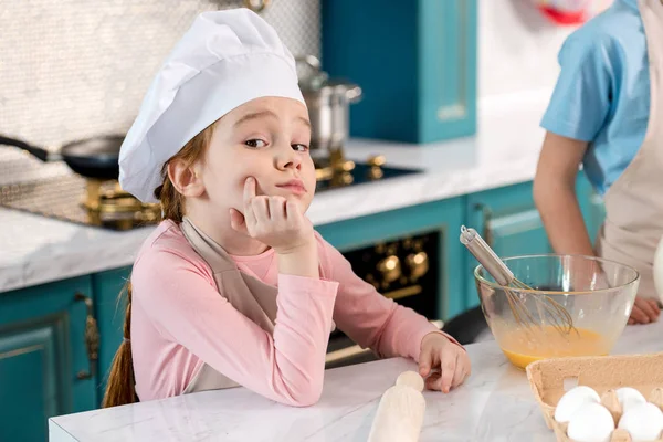 Niedliches Kind mit Kochmütze und Schürze sitzt mit der Hand am Kinn und blickt in der Küche in die Kamera — Stockfoto