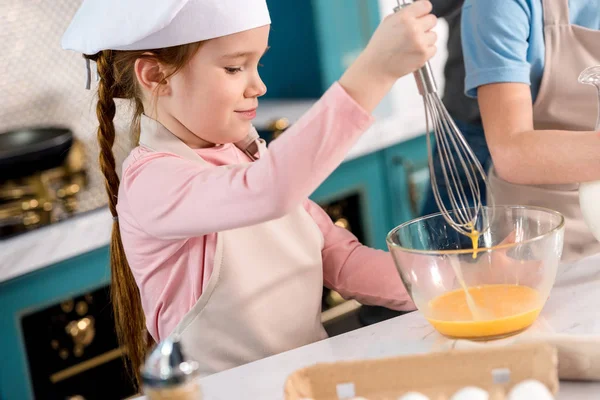 Ребенок в шляпе шеф-повара и фартуке взбивая яйца на кухне — стоковое фото