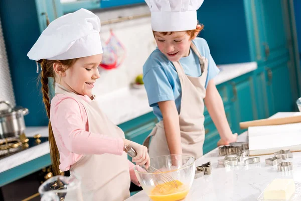 Чарівні усміхнені діти в капелюхах шеф-кухаря і фартухах роблять тісто разом на кухні — стокове фото