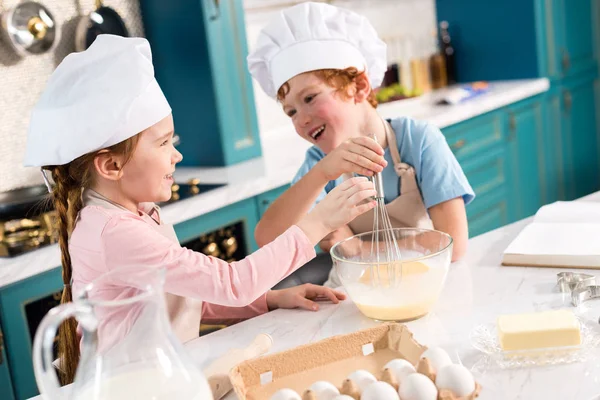 Mignons petits enfants dans les chapeaux de chef se souriant tout en fouettant la pâte dans la cuisine — Photo de stock