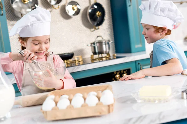 Fröhliche kleine Kinder in Kochmützen und Schürzen, die in der Küche Teig zubereiten — Stockfoto