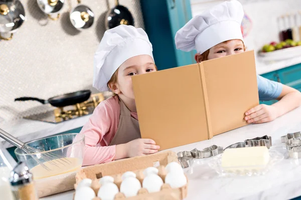 Entzückende Kinder mit Kochmützen halten Kochbuch in der Hand und blicken in die Kamera — Stockfoto