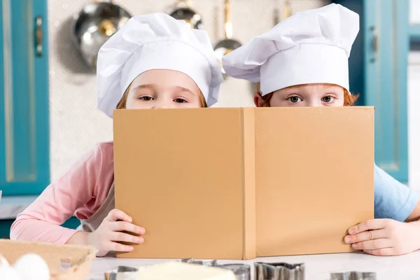 Niedliche Kinder mit Kochmützen halten Kochbuch in der Hand und blicken in die Kamera — Stockfoto