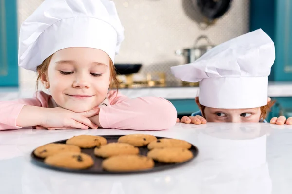 Adorables enfants heureux dans des chapeaux de chef en regardant les biscuits savoureux sur la table — Photo de stock