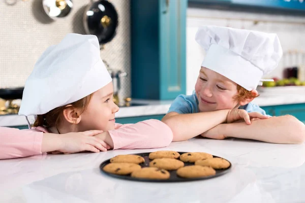 Niños felices en sombreros de chef sonriendo entre sí y deliciosas galletas en la mesa - foto de stock