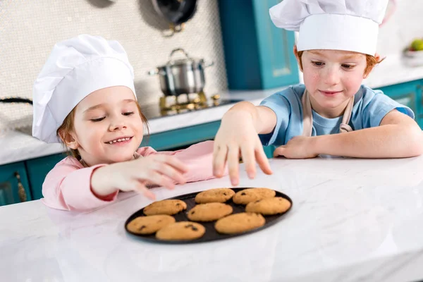 Niños felices en sombreros de chef comiendo galletas sabrosas en la cocina - foto de stock