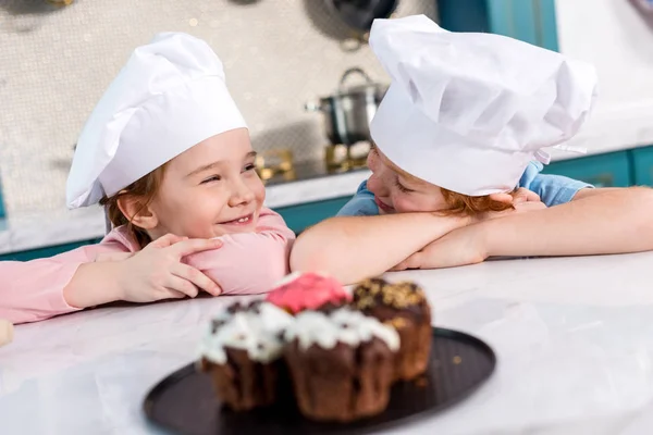 Adorables niños en sombreros de chef sonriendo entre sí y deliciosos cupcakes en primer plano - foto de stock