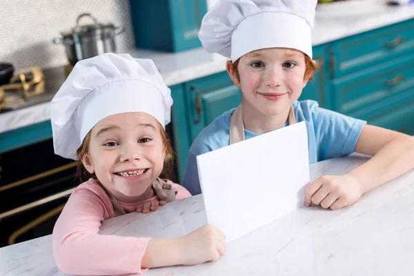 Carino bambini piccoli in cappelli da chef in possesso di carta bianca e sorridente alla fotocamera — Foto stock