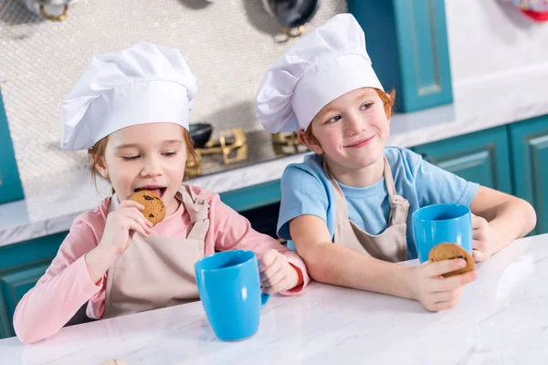 Bambini sorridenti in cuoco cappelli bere tè e mangiare biscotti in cucina — Foto stock