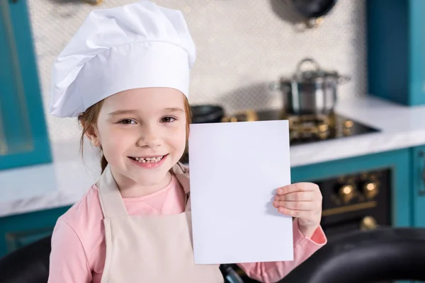 Nettes Kind mit Kochmütze und Kochschürze mit leerer Karte und lächelnd in die Kamera — Stockfoto