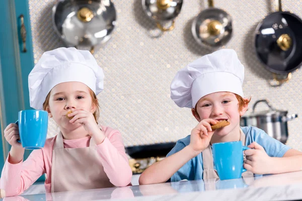 Niños con galletas y tazas de té sonriendo a la cámara en la cocina - foto de stock