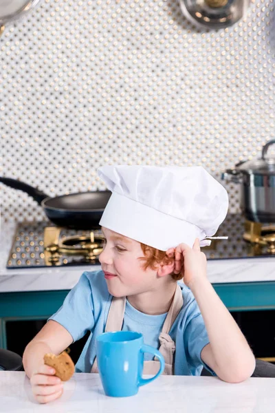 Милий маленький хлопчик у капелюсі шеф-кухаря п'є чай і їсть печиво на кухні — стокове фото