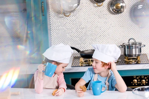 Милые детишки в шляпах шеф-повара пьют чай и едят печенье на кухне — стоковое фото