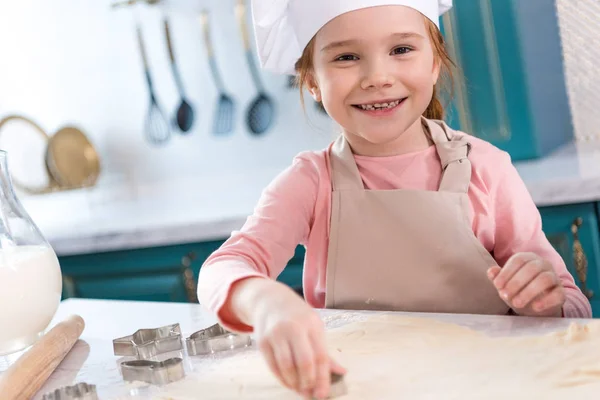 Adorable child in chef hat and apron smiling at camera while preparing cookies — Stock Photo