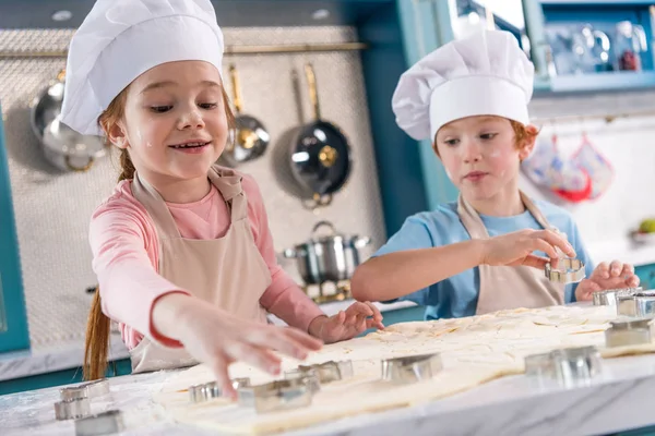 Чарівні маленькі діти в капелюхах шеф-кухаря і фартухах готують печиво разом — стокове фото