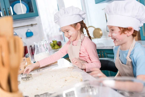 Niños felices en sombreros de chef preparando galletas juntos en la cocina - foto de stock