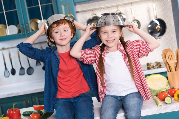 Entzückende kleine Kinder mit Utensilien auf dem Kopf, die in der Küche in die Kamera lächeln — Stockfoto