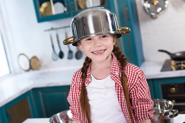 Criança adorável com panela na cabeça sorrindo para a câmera na cozinha — Fotografia de Stock