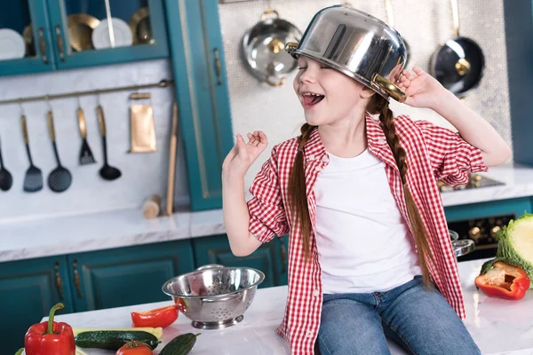 Bambino felice con padella sulla testa seduto sul tavolo della cucina — Foto stock