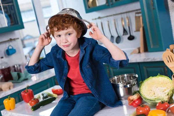 Netter kleiner Junge mit Pfanne auf dem Kopf lächelt in die Kamera, während er auf dem Küchentisch sitzt — Stockfoto