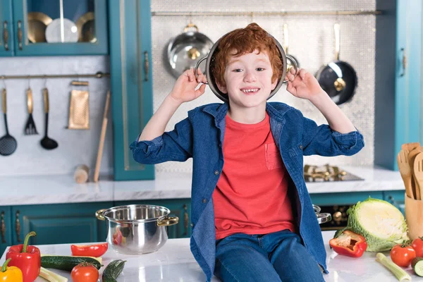 Adorable little boy with pan on head smiling at camera while sitting on kitchen table — Stock Photo