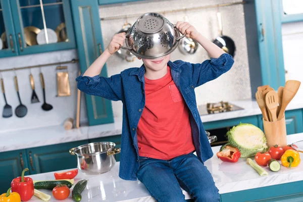 Lindo niño pequeño con sartén en la cabeza sentado en la mesa de la cocina — Stock Photo