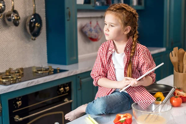 Kid holding digital tablet and looking away while sitting on kitchen table — Stock Photo