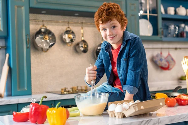 Süßer kleiner Junge lächelt in die Kamera, während er in der Küche kocht — Stockfoto