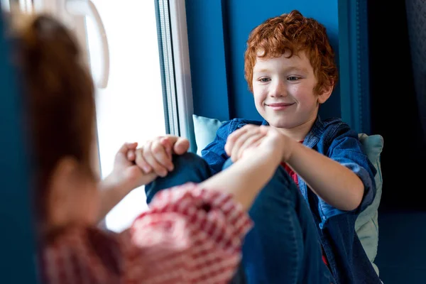 Recortado disparo de lindos niños pequeños cogidos de la mano y sonriendo entre sí mientras se sienta en el alféizar de la ventana - foto de stock