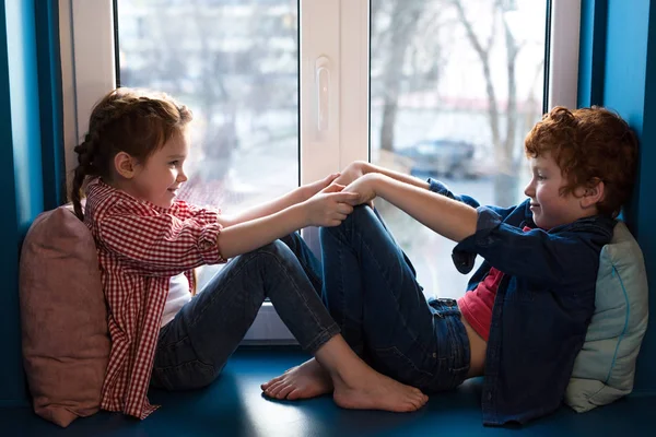 Entzückende kleine Kinder halten Händchen und lächeln einander an, während sie auf der Fensterbank sitzen — Stockfoto