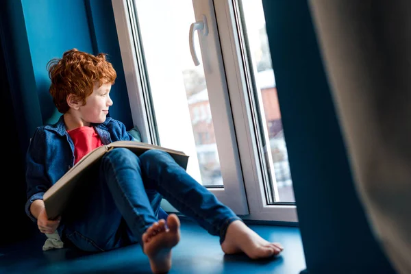 Carino ragazzo a piedi nudi con libro seduto sul davanzale della finestra — Foto stock