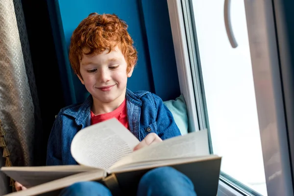 Netter lächelnder kleiner Junge liest Buch, während er auf der Fensterbank sitzt — Stockfoto