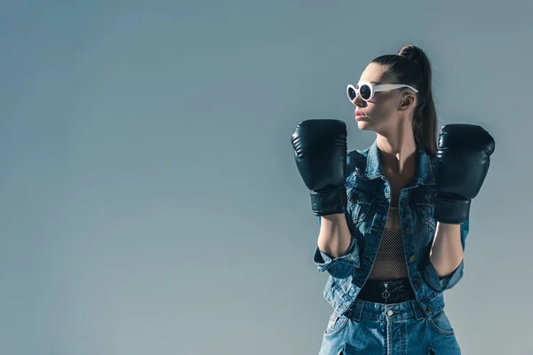 Стильная девушка в джинсовой одежде и солнцезащитных очках позирует в боксёрских перчатках, изолированная на сером — стоковое фото
