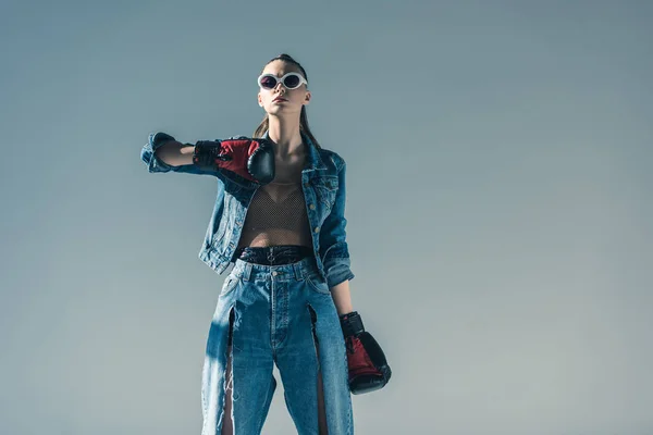 Belle fille élégante en denim et lunettes de soleil posant avec des gants de boxe, isolé sur gris — Photo de stock