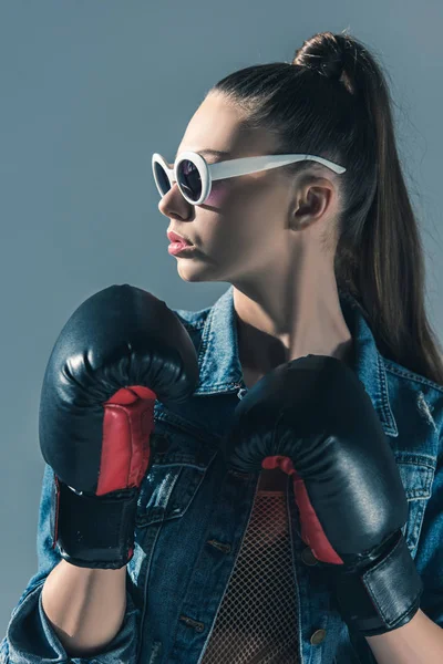 Chica con estilo en denim y gafas de sol posando con guantes de boxeo, aislado en gris - foto de stock