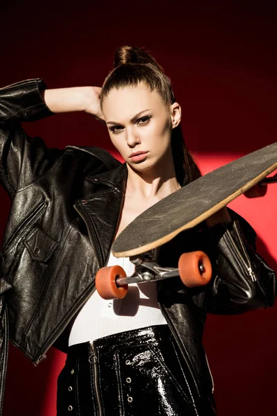 Attractive female skater in black leather jacket posing with longboard, on red — Stock Photo