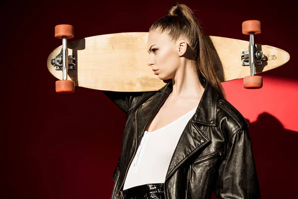 Beautiful girl in trendy leather jacket posing with longboard, on red — Stock Photo