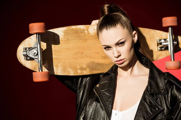 Attractive fashionable girl in black leather jacket posing with longboard, on red — Stock Photo