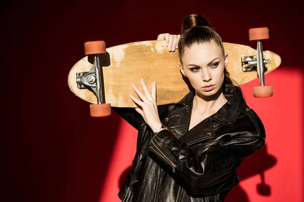 Attractive girl in black leather jacket posing with longboard, on red — Stock Photo
