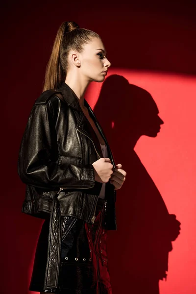 Vue latérale de la fille élégante avec coiffure en queue de cheval posant en veste en cuir noir pour le tournage de mode sur rouge — Photo de stock