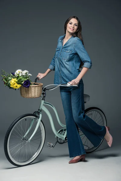 Hermosa chica en ropa de mezclilla sosteniendo bicicleta con flores en canasta de mimbre, en gris - foto de stock