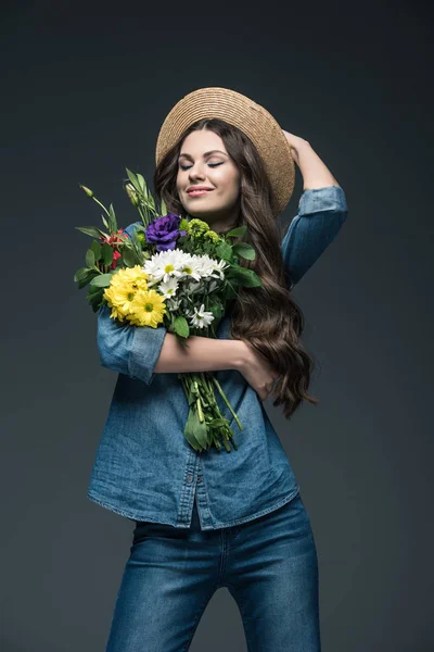 Atractiva chica sonriente con los ojos cerrados en ropa de mezclilla y sombrero de paja con ramo de flores, aislado en gris - foto de stock