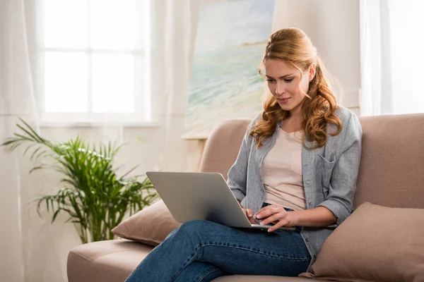 Bela jovem mulher usando laptop enquanto sentado no sofá em casa — Fotografia de Stock