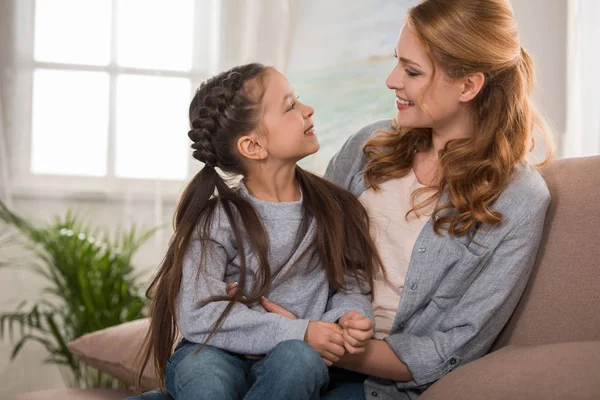 Glückliche Mutter und Tochter lächeln einander an, während sie zu Hause zusammen auf dem Sofa sitzen — Stockfoto