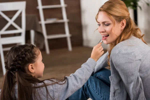 Carino piccola figlia applicando rossetto per felice madre a casa — Foto stock