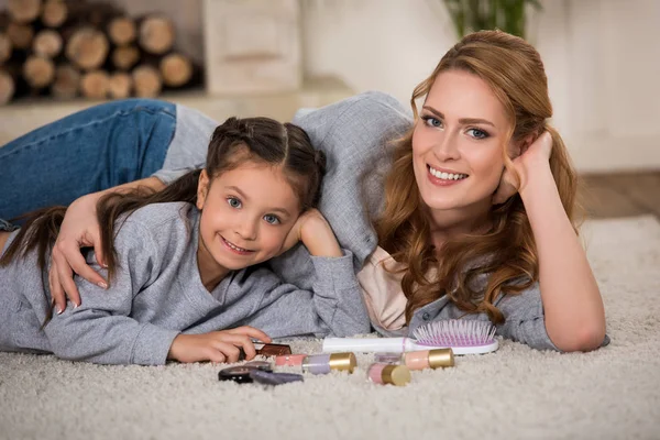 Happy mother and daughter lying on carpet with cosmetics and smiling at camera — Stock Photo