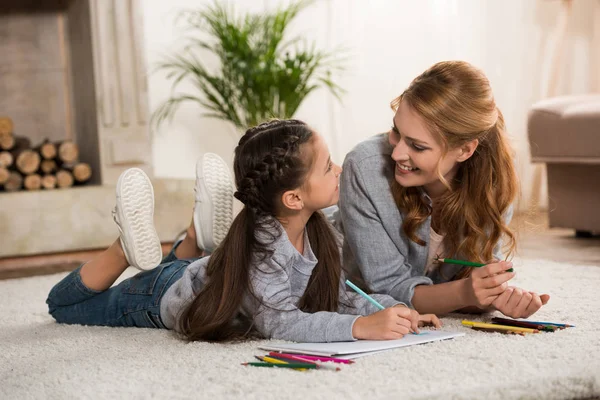 Feliz madre e hija pequeña dibujo con lápices de colores y sonriendo el uno al otro en casa - foto de stock